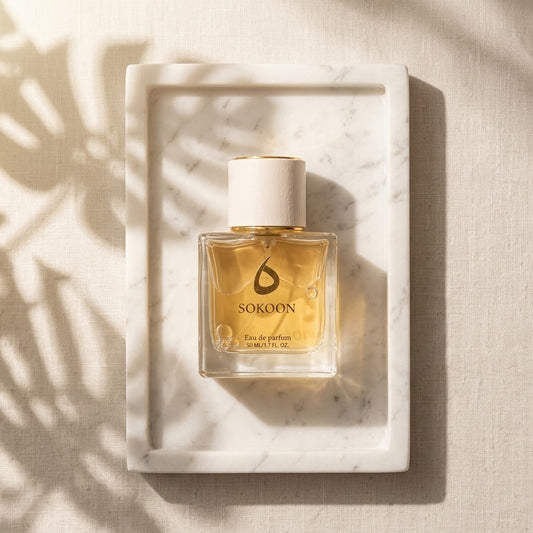A bottle of SOKOON Eau de Parfum with golden liquid and a white cap, presented in a white marble tray on a textured beige linen background with soft leaf shadows.